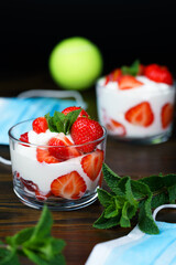 Whipped cream and strawberries served in a glass. Dark wooden table, tennis ball in bokeh, surgical masks, high resolution