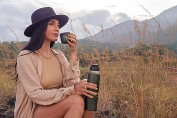 A young woman drinks tea from a thermos against the backdrop of the savannah and mountains. Safari style in clothing. Travel, camping.