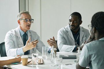 Portrait of mature doctor sitting at meeting table in conference room while speaking to people...