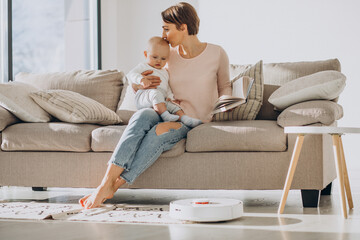 Young mother sitting on sofa with toddler son and watching robot vacuum cleaner doing housework