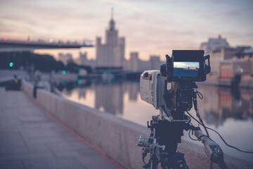 A professional video camera stands on a tripod recording the city and the river at sunrise,...