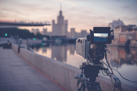 A professional video camera stands on a tripod recording the city and the river at sunrise, documentary filming in Moscow