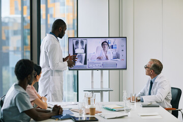 Obraz premium Portrait of African-American doctor standing by digital screen in onference room and talking to expert via video call, copy space