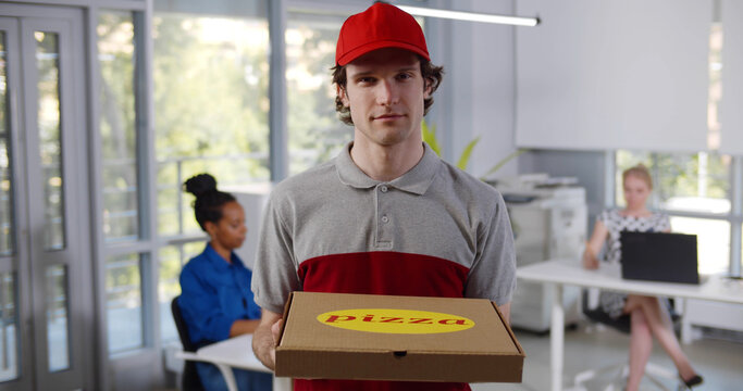 Portrait of delivery guy in uniform holding pizza box, looking at camera and talking to client in modern office