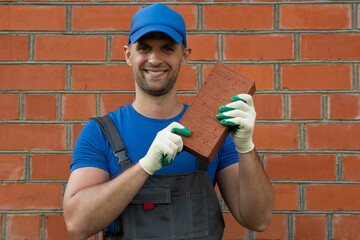 A man wearing a cap and gloves shows a brick to the camera in a construction concept