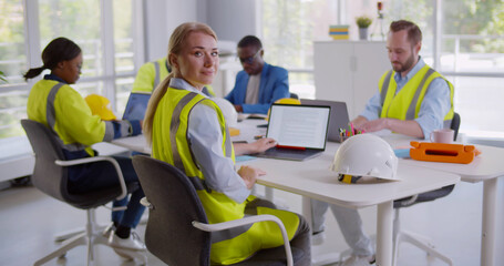 Young woman constructor wearing reflective vest posing at camera sitting at desk in modern office