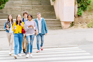 Obraz premium Full height of happy young group of five males and females students on a walk on a day off. A happy friends crossing the road at a pedestrian crossing. Front view.