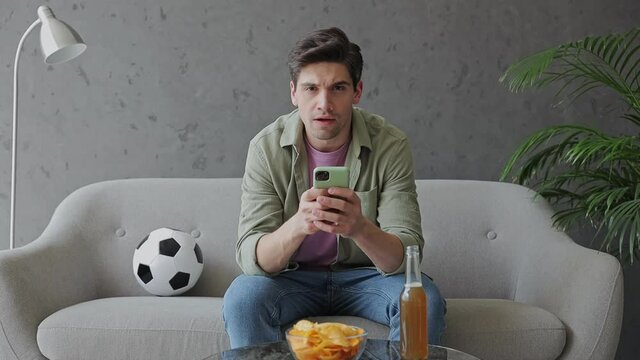 Young sad man fan in green clothes t-shirt cheer up support sport football team sit on grey sofa with soccer ball use toss cell mobile phone bottle beer ale eat chips rest watch tv indoors room gray