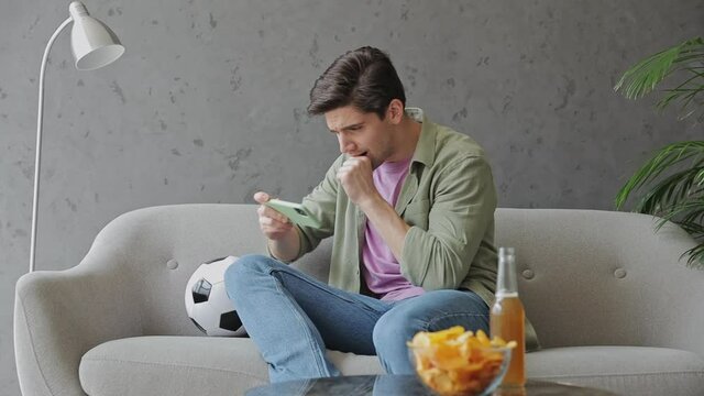 Young sad upset man fan in green clothes t-shirt cheer up support sport football team sit on grey sofa with soccer ball bottle beer eat chips wait watch tv on mobile cell phone indoors room gray wall