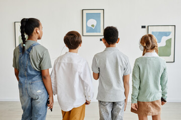 Minimal back view at multi-ethnic group of children looking at paintings in modern art gallery and wearing headphones - obrazy, fototapety, plakaty
