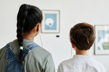 Back view at two teenage kids looking at paintings in modern art gallery and wearing headphones