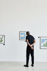 Minimal full length back view at African-American man looking at paintings in modern art gallery, copy space