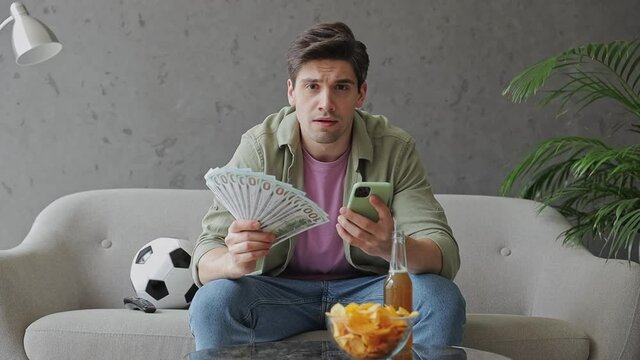 Young fun happy man fan in green clothes t-shirt cheer up support sport football team sit on grey sofa with soccer ball bottle beer eat chips wait watch tv hold money mobile cell phone win indoor room