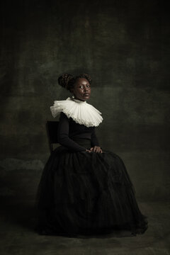 Medieval African young woman in black vintage dress with big white collar posing isolated on dark green background. Concept of comparison of eras, modernity and renaissance.