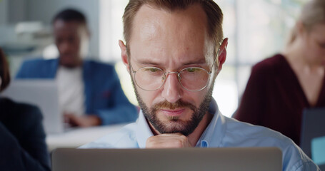 Fototapeta premium Close up of young businessman sitting at desk working on laptop and looking thoughtfully in office.