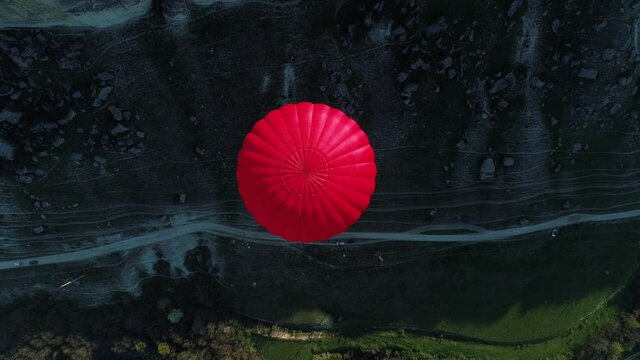Aerial top view of a red hot air balloon floating over a mountain slope, a narrow path, and a green meadow. Shot. Concept of travelling and adventure.