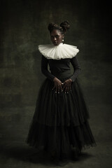 Medieval African young woman in black vintage dress with big white collar posing isolated on dark green background. Concept of comparison of eras, modernity and renaissance.