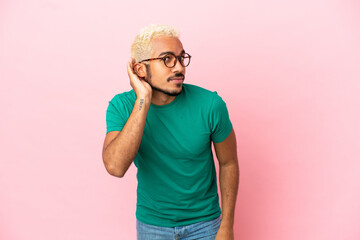 Young Colombian handsome man isolated on pink background listening to something by putting hand on the ear