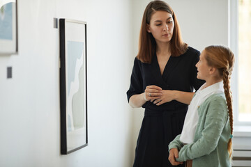 Side view portrait of mother and daughter visiting modern art gallery together and looking at...