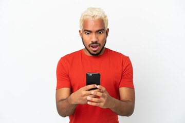 Fototapeta premium Young Colombian handsome man isolated on white background looking at the camera while using the mobile with surprised expression