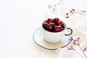 Raw sweet cherries in a cup and saucer and white tablecloth with wildflowers, copy space.