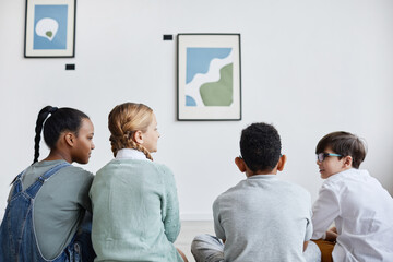 Minimal back view at diverse group of children sitting on floor in modern art gallery and looking at paintings, copy space