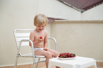 White child eats strawberries in summer sitting on the balcony on the background of the roofs