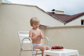 White kid eats strawberries in summer sitting on the terrace on the background of the roofs