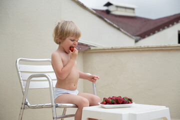 Boy eats red strawberries in summer sitting on the terrace on the background of the roofs