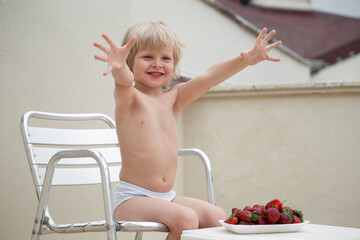 Happy boy eats strawberries in summer sitting on the terrace on the background of the roofs