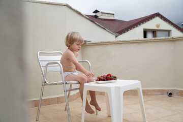 White child eats strawberries in summer sitting on the veranda on the background of the roofs