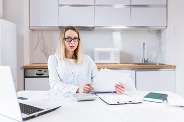 young female housewife sitting a table in house kitchen an incomprehensible deal with paperwork bills and loans. Woman is exhausted and frustrated with documents of enterprise or family budget at home