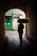 The silhouette of a girl with an umbrella in the arch, looking away from the camera, goes into a small courtyard. Walking down the street with an umbrella