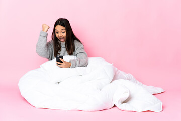 Young mixed race woman wearing pijama sitting on the floor with phone in victory position