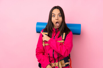 Young mountaineer girl with a big backpack over isolated pink background surprised and pointing side