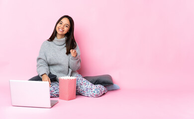 Young mixed race woman eating popcorn while watching a movie on the laptop pointing front with happy expression