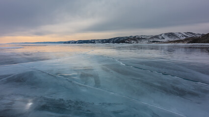 Sunset over the endless frozen lake. Cracks and reflections of the setting sun on the smooth shiny surface of the ice. Snow-covered mountain range against the backdrop of a cloudy evening sky. Baikal