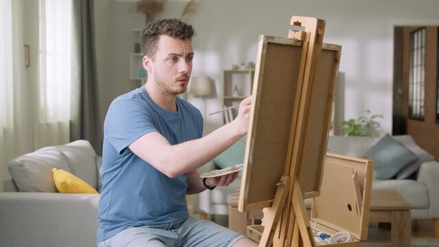 Young man is working in his modern home studio in the morning, drawing on canvas, enjoying the process of creating new artwork, inspired by natural beauty, Slow motion.