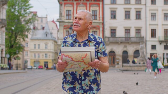Elderly tourist stylish grandfather getting lost in big city trying to find route. Excited senior traveler man walking along street, looking for way using paper map in old town. Summer vacation trip