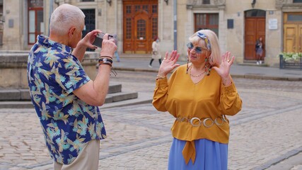Front view of senior couple man and woman tourists. Elderly grandfather taking photo pictures of grandmother on retro camera. Family enjoying vacations time together in summer old town Lviv, Ukraine