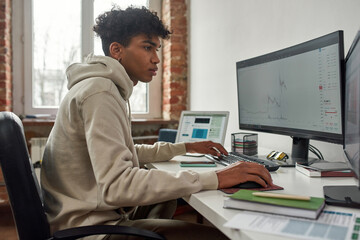 Focused young man looking at pc screen while trading online using computer, sitting at home in modern living room office