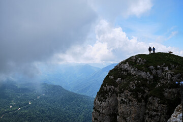 Silhouettes of two travelers, man, woman and dog on top of cliff. Couple of tourist people are standing on top of mountain together against background of mountains and fog.