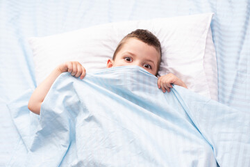 A frightened four-year-old toddler boy covered his face with a blue blanket. Peek out from under the covers.