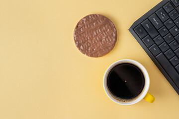 Chocolate cake and coffee nearby keyboard on yellow background. A cup of black coffee or chicory drink and sweet crispy snack for lunch at work.
