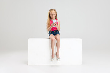 One beautiful little girl in casual clothes sitting on box isolated on white studio background....