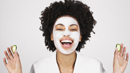 Cheerful african american woman in face mask holding slices of cucumber isolated on grey