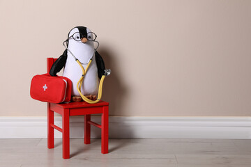 Toy penguin with eyeglasses, stethoscope and first aid bag near beige wall, space for text. Pediatrician practice © New Africa