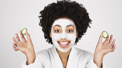 Smiling african american woman with mask on face holding slices of cucumber isolated on grey