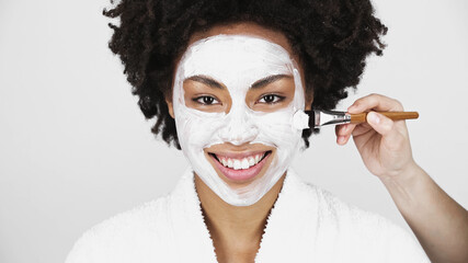 Man with brush applying mask on face of cheerful african american woman isolated on grey