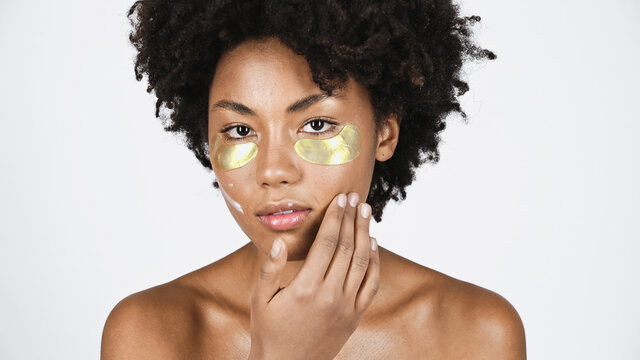 Young African American Woman With Golden Eye Patches Applying Cosmetic Cream Isolated On Grey